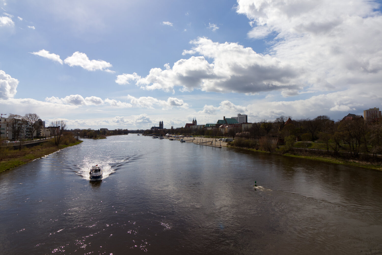 Wide view of a river with buildings on both sides. A boat approaches the camera.