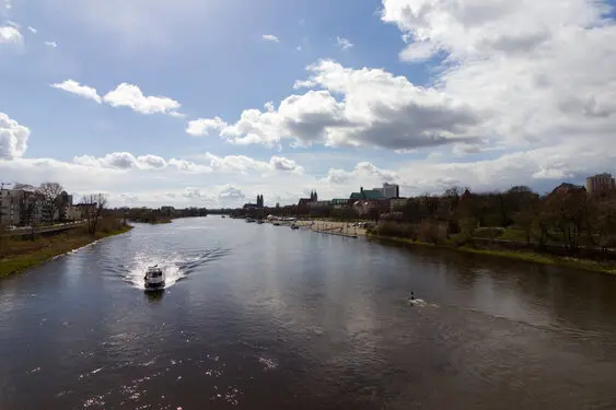 Wide view of a river with buildings on both sides. A boat approaches the camera.