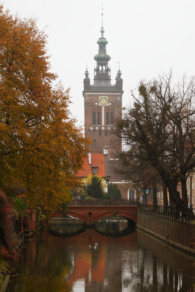 Small river with a bridge and a brick tower building in the background. Autumn trees at the sides of the river.