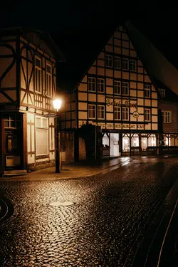 A dark alley illuminated by a street lamp at night. A cobblestone road is slightly wet, reflecting the light. Half-timbered buildings are in the background.