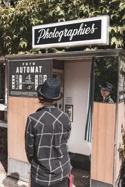 A French photo booth with a 'photographies' sign. A woman with a hat stands in front of it. Her face is reflected in a mirror attached to the photo booth.