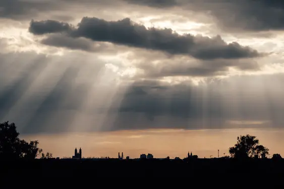 A cityscape silhouette with big clouds above it. Beams of sunlight come through the clouds, hitting the cityscape.