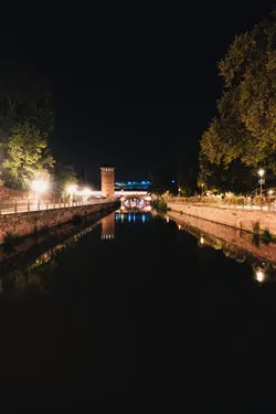 A water channel at night. Light lamps on both sides of the channel illuminate the sidewalks. A blue light shines in the background and is reflected in the water. A few stars are visible in the sky.