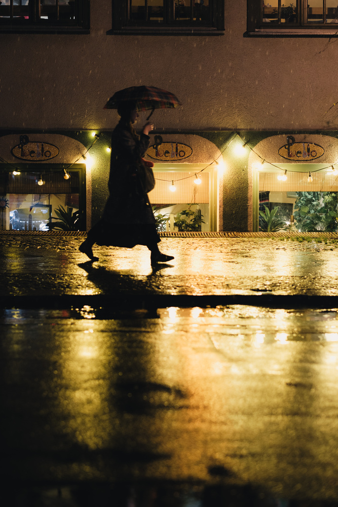 A woman holding an umbrella walks on a sidewalk at night. A chain of lightbulbs illuminates the scene.