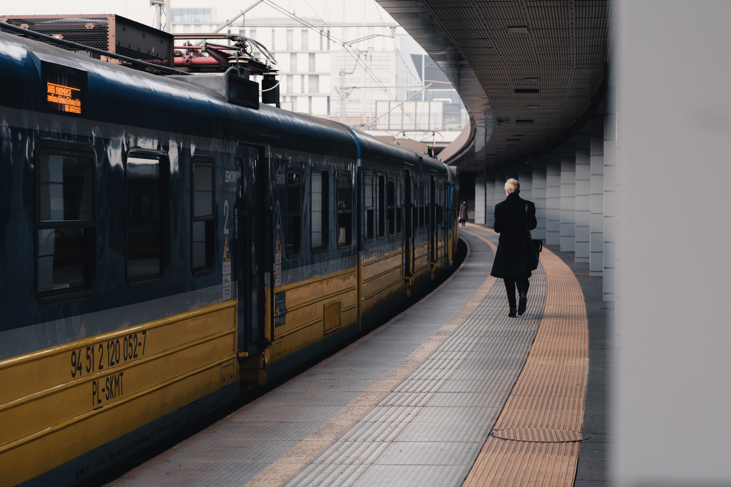 A curved train station. A blue/yellow train stands on the left side. A person with dark clothes and short blond hair walks away from the camera. The pillars of the train station form a curve.