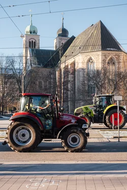 A green and red tractors on a city street, going in opposite directions. Tractor drivers look at each other. A church in the background.