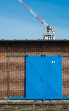 A blue metal gate in front of a red brick wall. Number 13 on the gate. A white-red crane above.