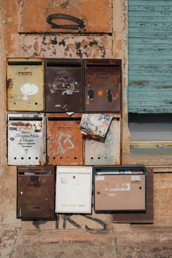 Nine rusty, decayed mail boxes. Each box has a different color. Partially visible window, covered by teal wood.