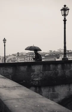 Monochrome photograph. A man with an umbrella walks on a stone bridge. A lamp post is on either side of the person.