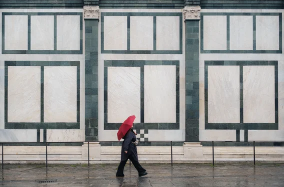 A woman with a red umbrella walks in front of a big marble wall with rectancle patterns.