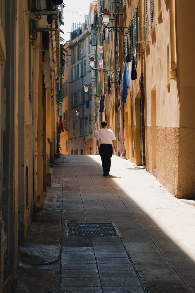 A man with a white t-shirt and black trousers walks through a mediterranean alley on a line where sunlight and shadow meet.