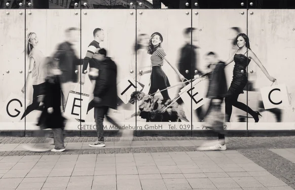 Motioned-blurred people walking in front of an advertising wall. A mix of sharp and motion-blurred people on the advertisement. The people on the ad hold shopping bags with the letters G, E, T, E and C.