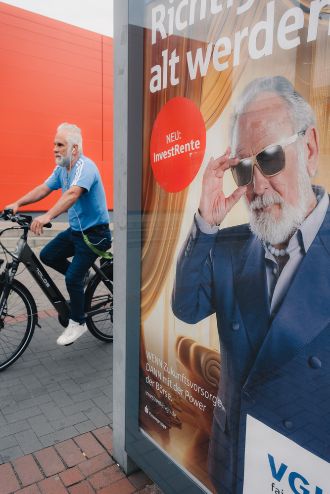 An advertisement wall shows an old man with gray hair and a beard. Next to the ad, there is a cyclist who matches the man from the ad visually.