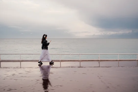A woman with a black jacket and a white maxi skirt walks from left to right. There is a reflection of her silhouette on the ground. A sea and clouds are in the background.