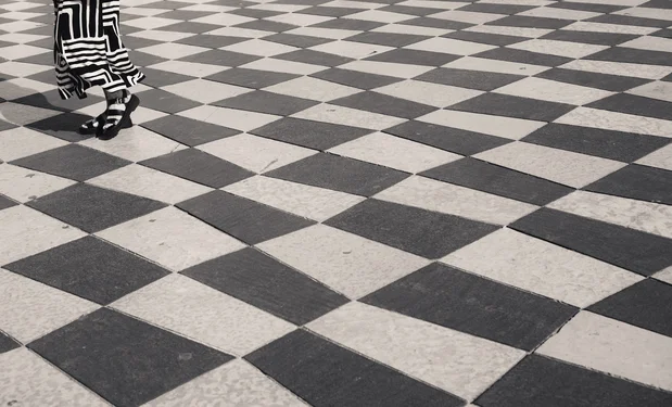 A person in the left upper corner wearing a black&white dress walks on a black&white check pattern floor. Only the dress and feet of the person are in view.