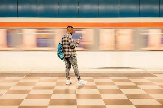 A man with a check pattern jacket standing on a check pattern floor, filming a moving subway with his phone.