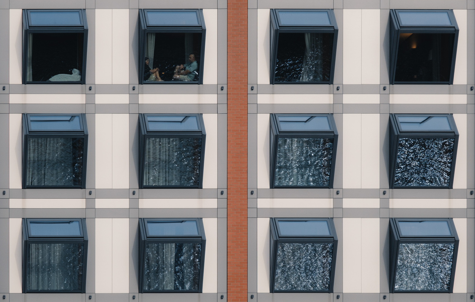 Twelve tilted windows on a building in a symmetrical composition. Some windows reflect water, which looks like broken glass. Two people sitting within one of the windows.