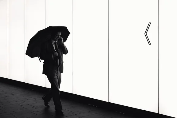 A man wearing a suit holds an umbrella and a bottle while having a phone call. He walks in front of a bright white background.