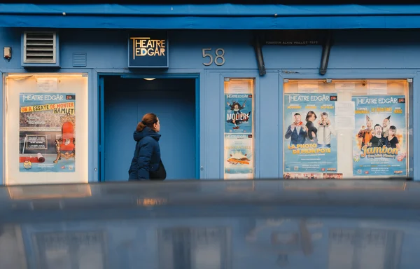 A woman wearing a blue jacket walks in front of a blue Theatre Edgar building. Blue posters with golden light hang on the building wall. The scene is reflected on a blue car roof.