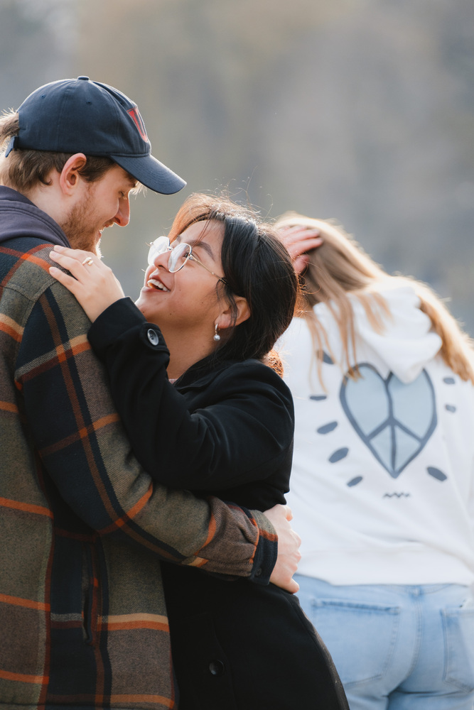 A hugging couple, looking into each other's eyes and smiling. A person in the background wears a hoodie with a mix of a heart and a peace symbol.