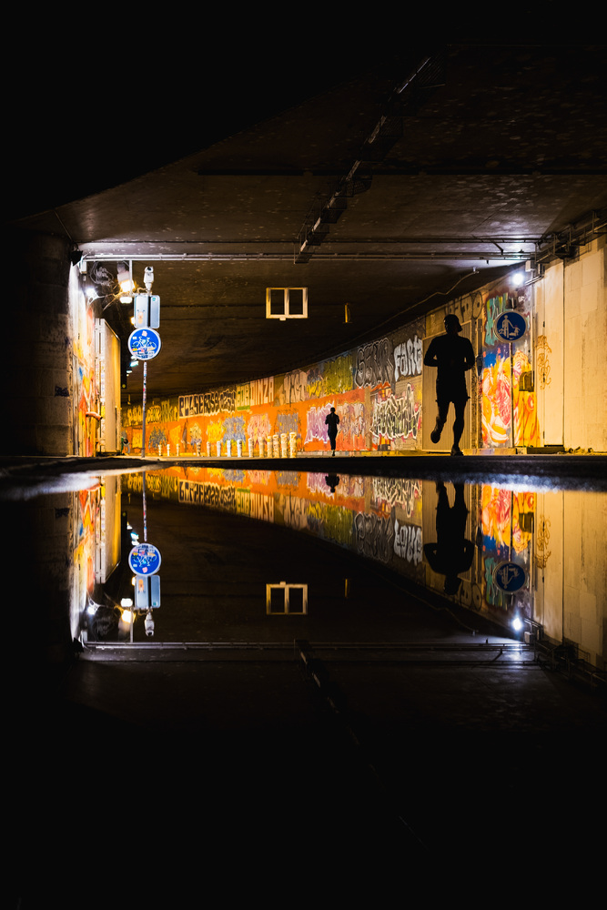 An entrance to a tunnel full of graffitis at night. Silhouettes of two joggers are visible. The scene is reflected in a puddle.